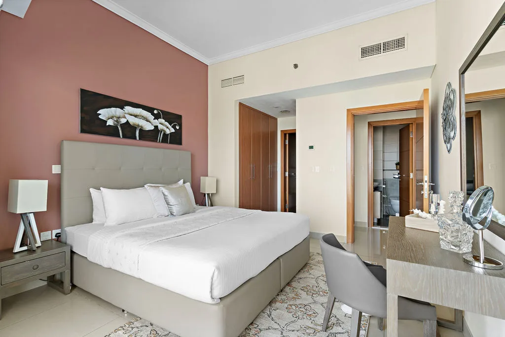 Un dormitorio con una cama grande, ropa de cama blanca y una pared rosa apagado.