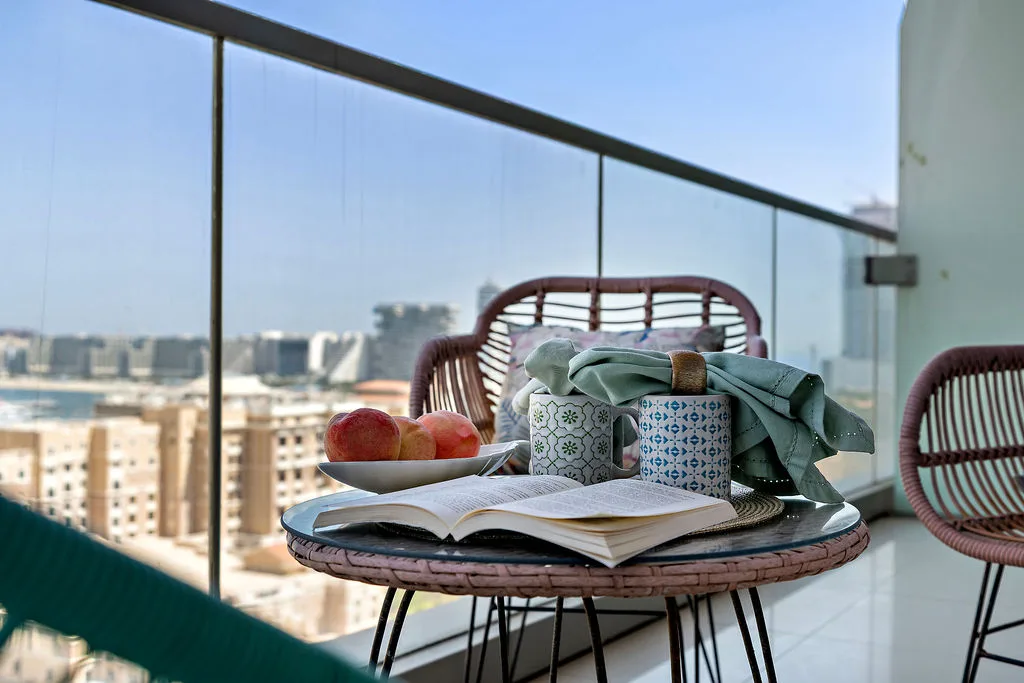 Una zona de estar en un balcón con un libro abierto, tazas, fruta y vistas a la ciudad.