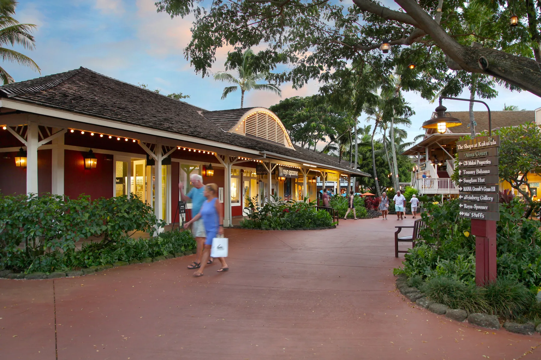 A paved walkway passes storefronts and landscaped greenery beneath large trees.