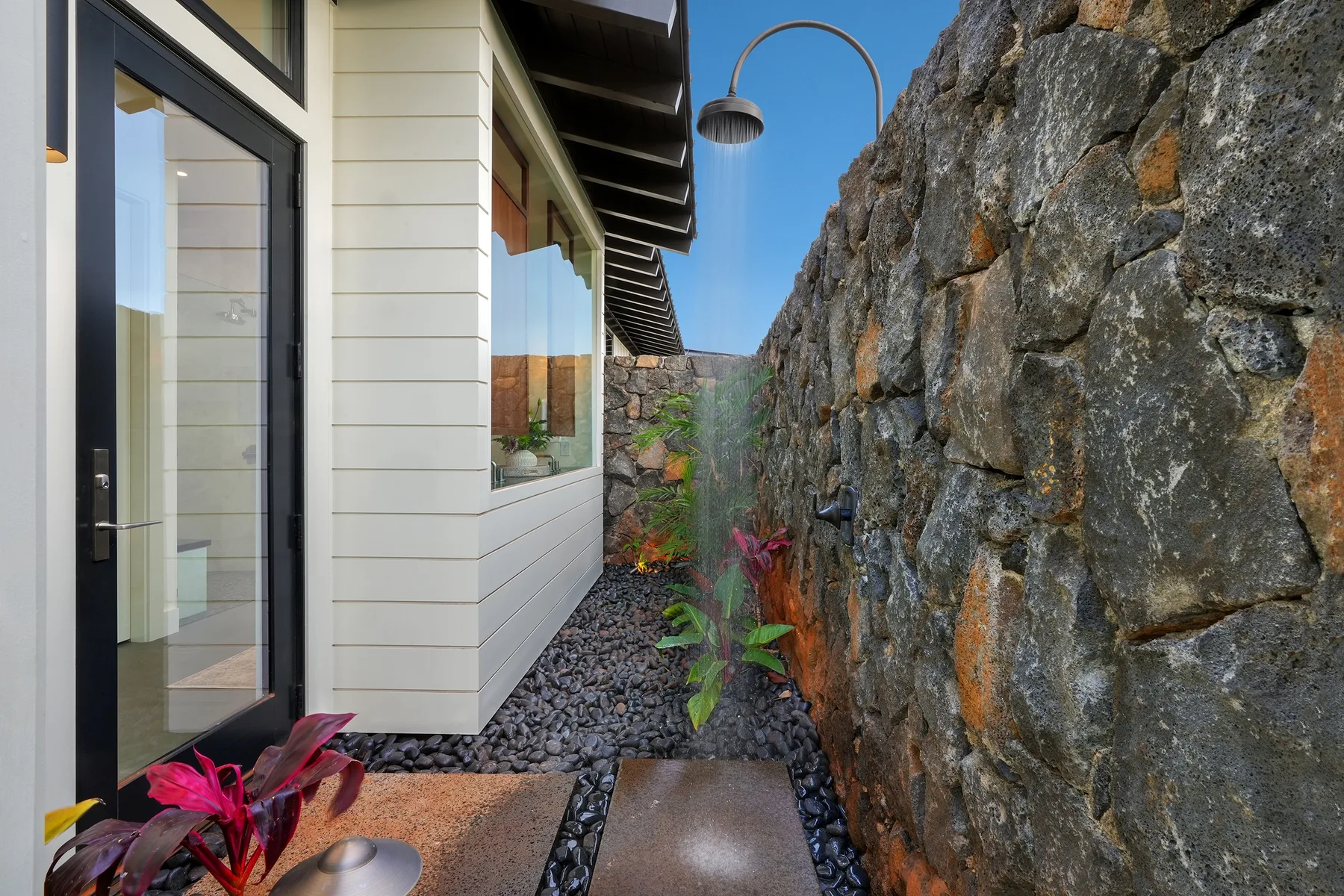 A narrow outdoor passage runs between a building and a stone wall, with plants and an overhead shower.