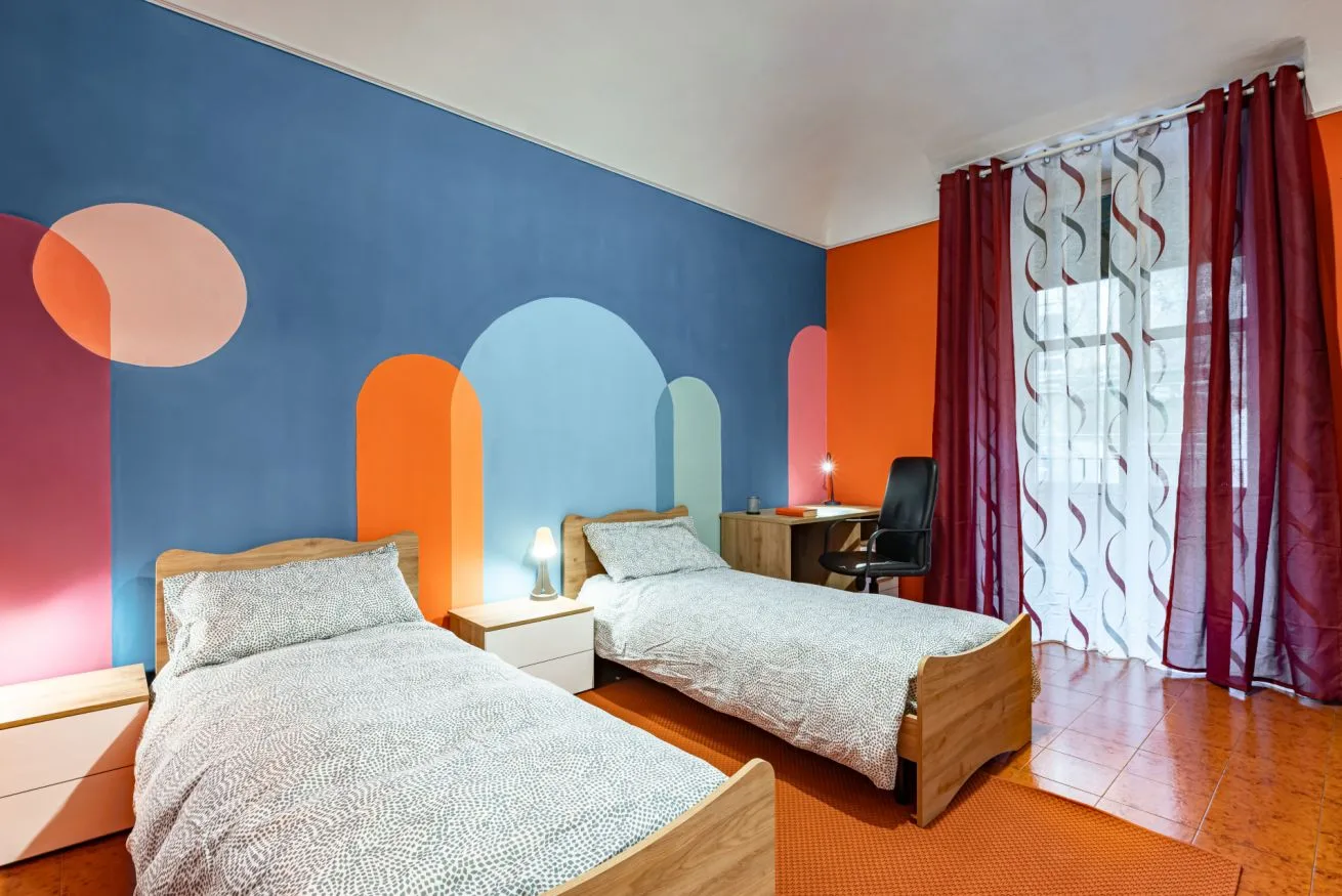 A colorful bedroom with two beds and a window framed by red curtains.