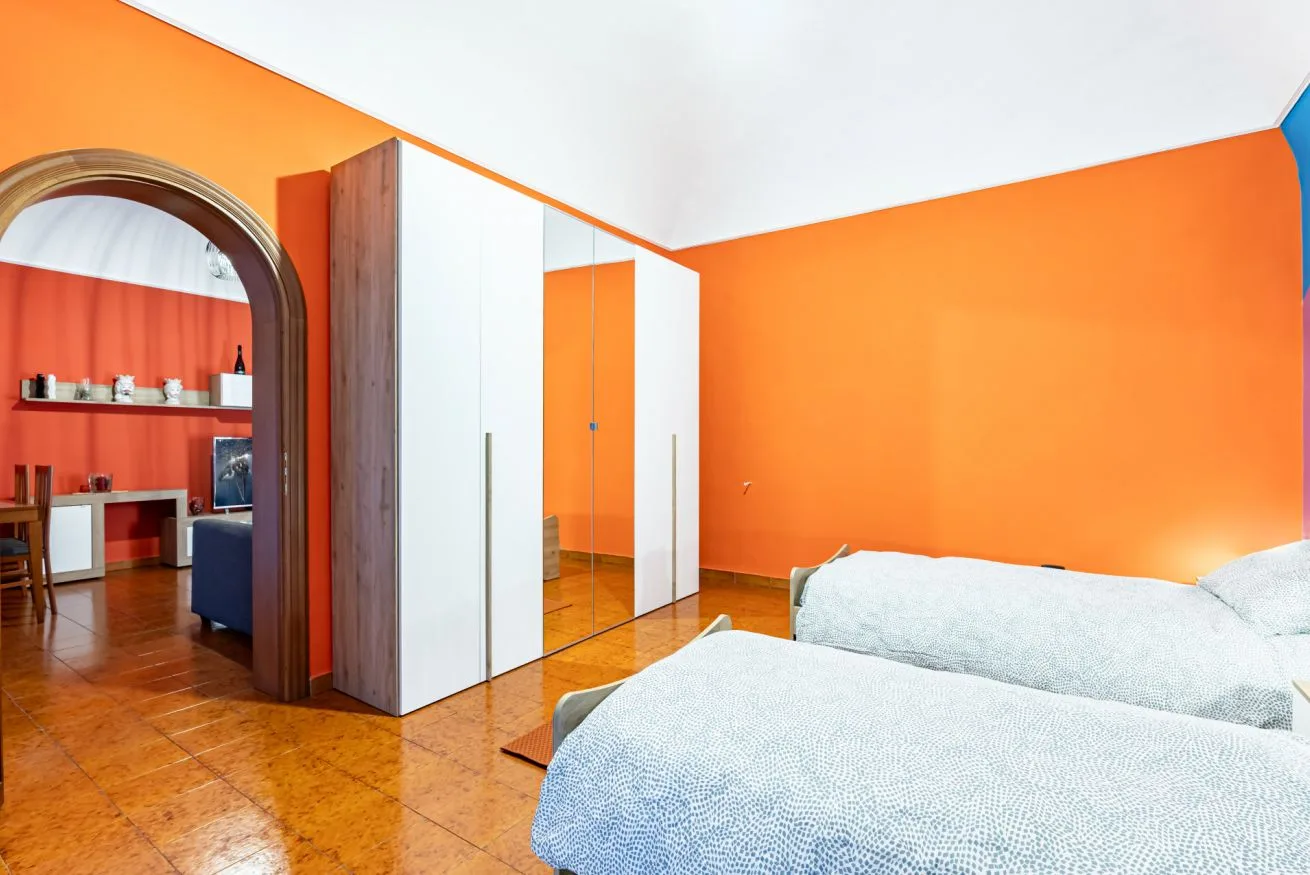 A bedroom with white bedding, mirrored wardrobes, and bright orange walls.