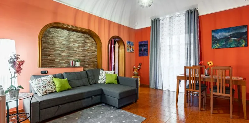 A living and dining area with a gray sectional, wooden table, and bright orange walls.