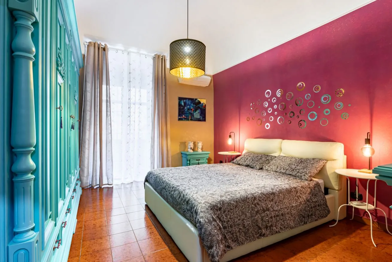 A bedroom with a bed against a magenta wall and turquoise cabinetry.