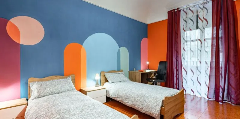 A colorful bedroom with two beds, bedside tables, and a desk by the window.