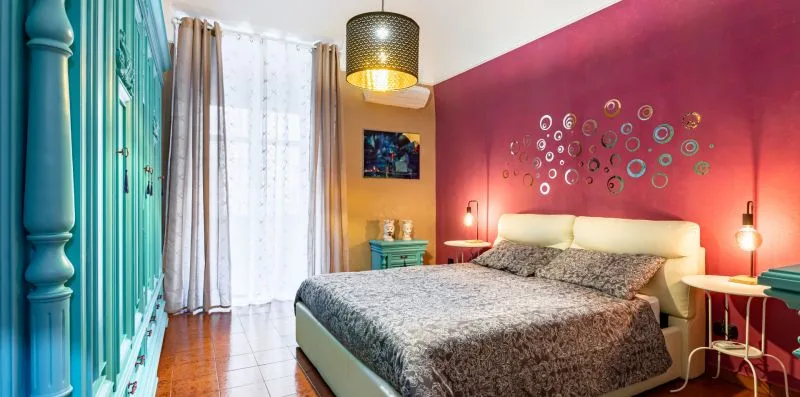 A bedroom with a double bed, colorful wall decorations, and tall windows.