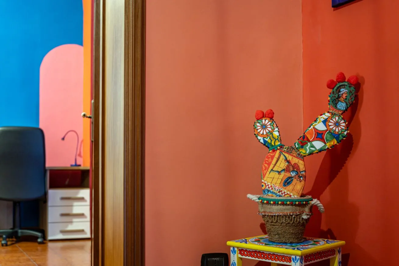 A colorful seated figure sculpture stands on a small decorated table against a red wall.