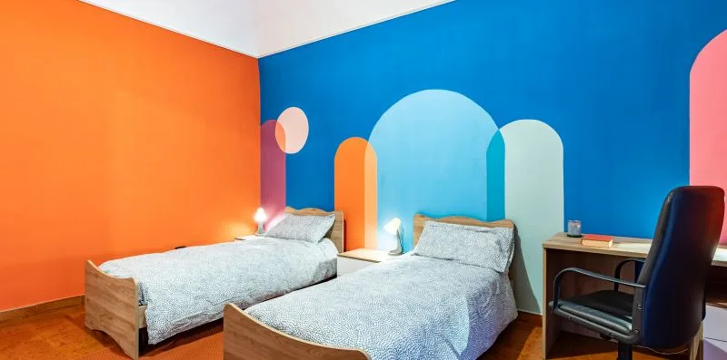 A colorful bedroom with two beds and a desk against a bright blue wall.
