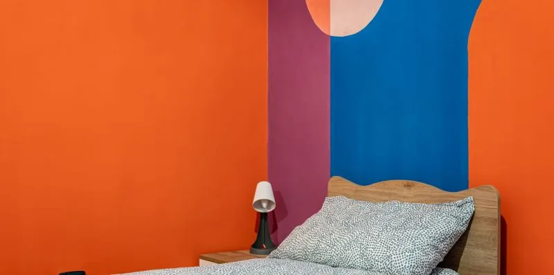 A bedroom with a bed against a bright orange wall and colorful mural.