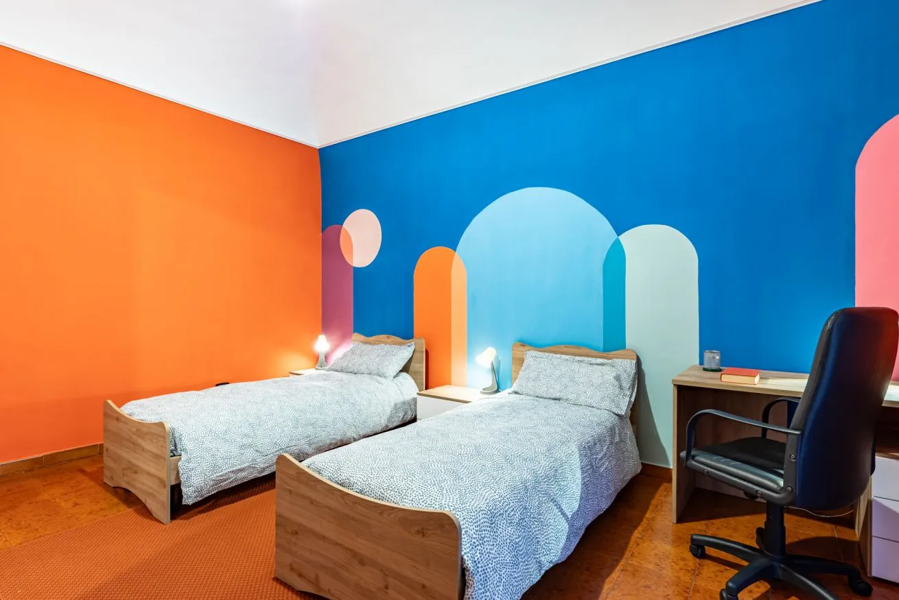 A colorful bedroom with two beds and a desk against a bright blue wall.