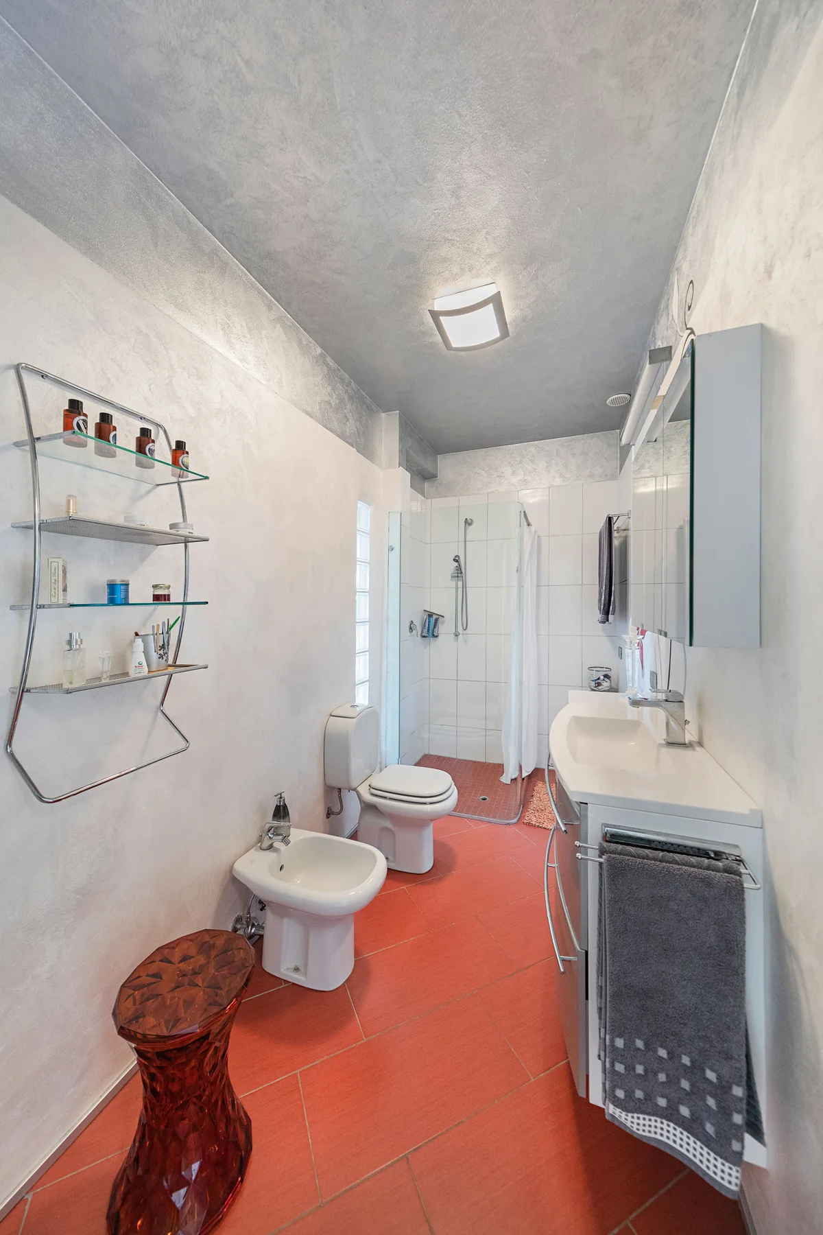 Un baño estrecho tiene ducha, inodoro y suelo de baldosas rojas.