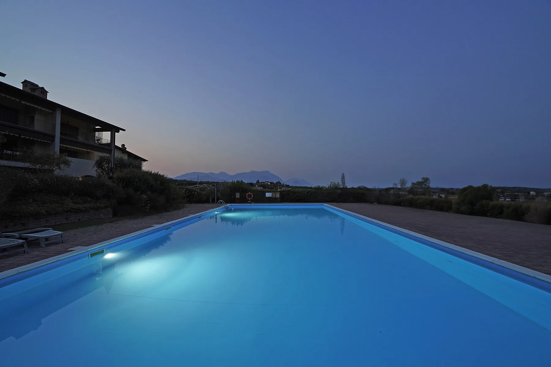 Una piscina exterior iluminada al anochecer, junto a una zona pavimentada.
