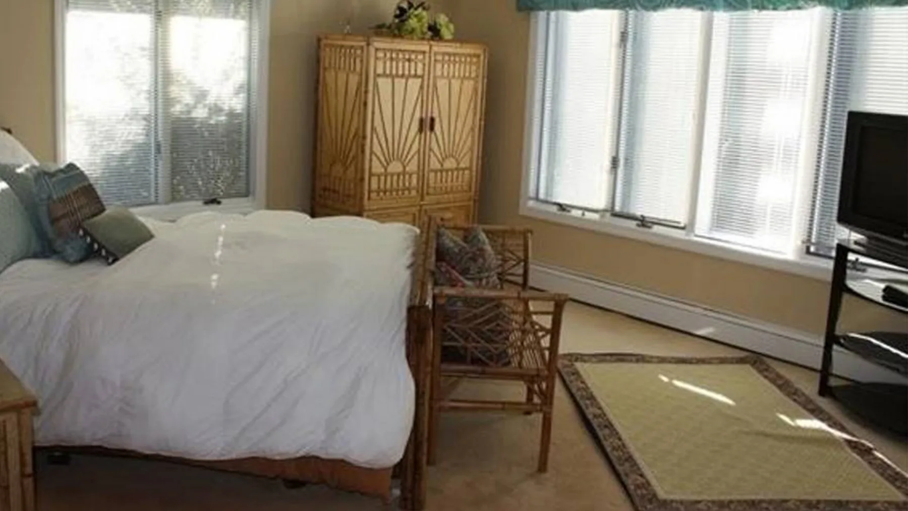 Un dormitorio con una cama, asientos de mimbre y un televisor junto a ventanas luminosas.
