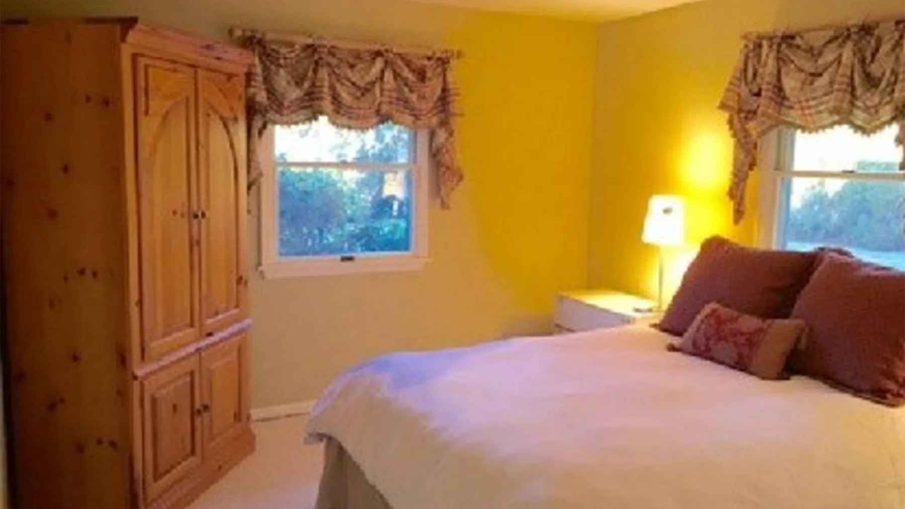 Un dormitorio con una cama, un armario de madera y dos ventanas con cortinas.