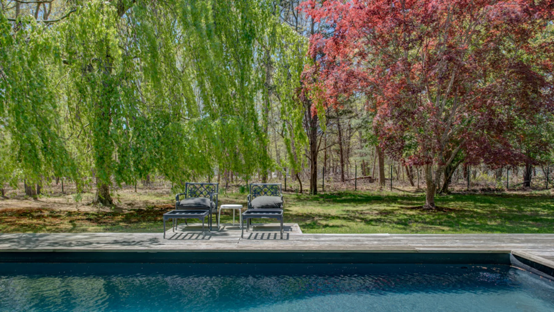 A swimming pool sits beside outdoor seating beneath leafy trees.