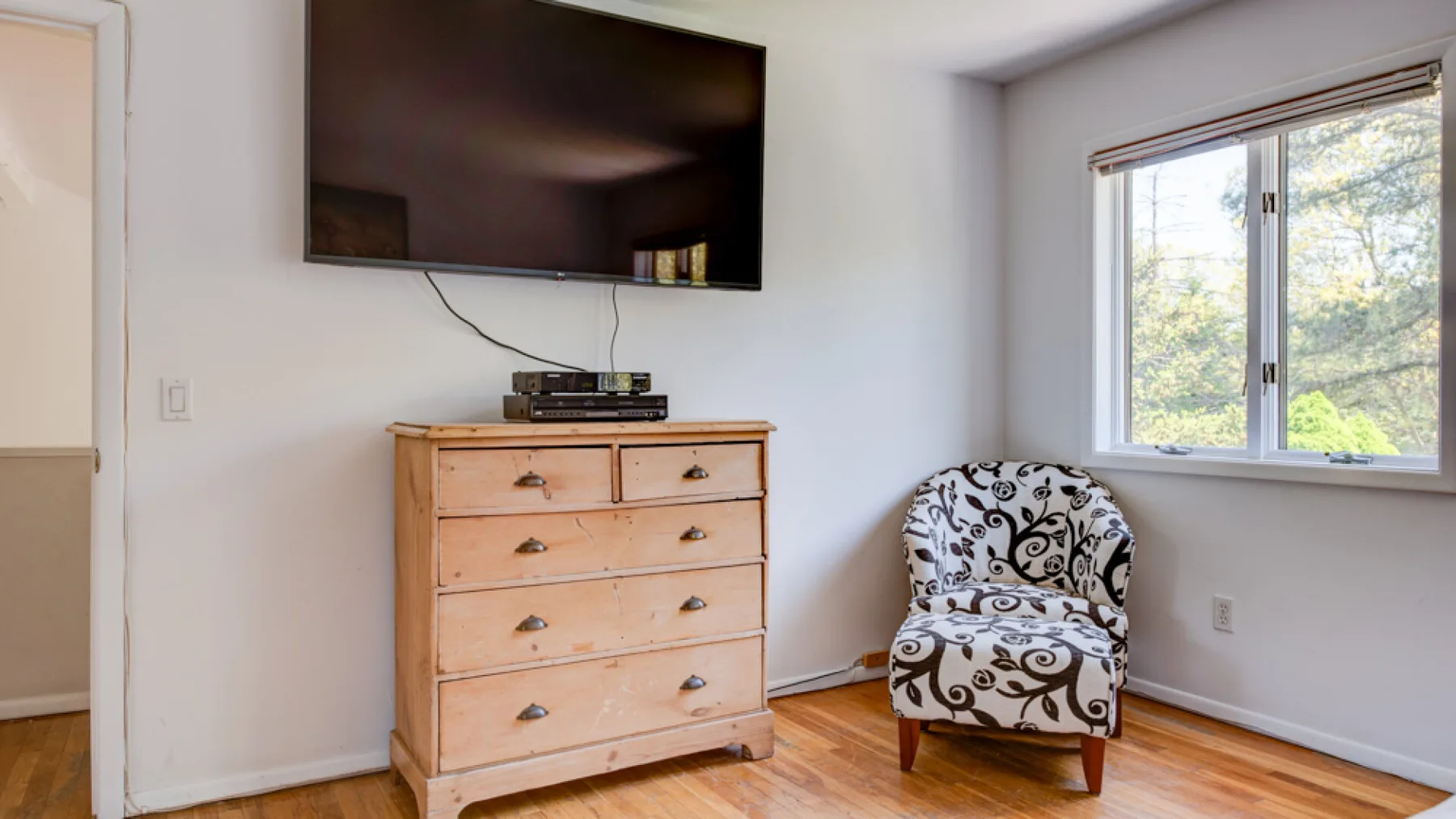 A bright bedroom with a wooden dresser, television, and patterned seating by the window.