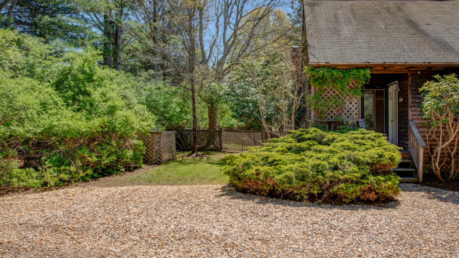 A gravel driveway leads to a wooden building, surrounded by shrubs and trees.