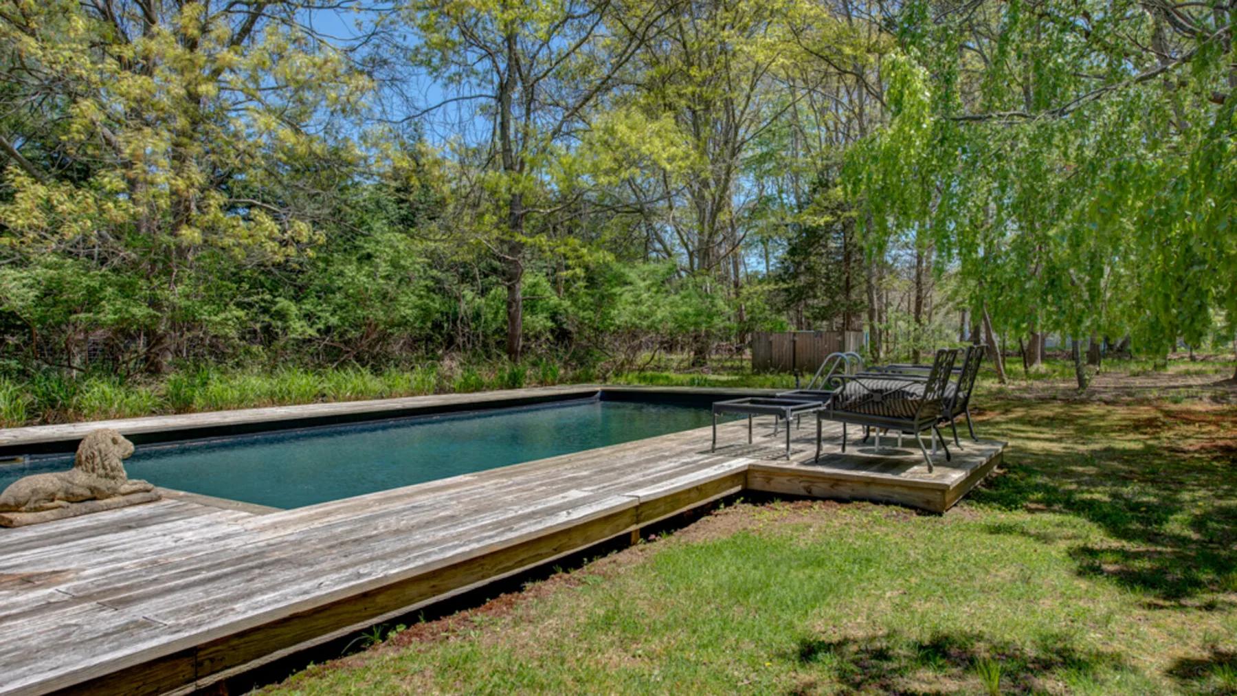 An outdoor swimming pool beside a wooden deck and seating in a leafy yard.