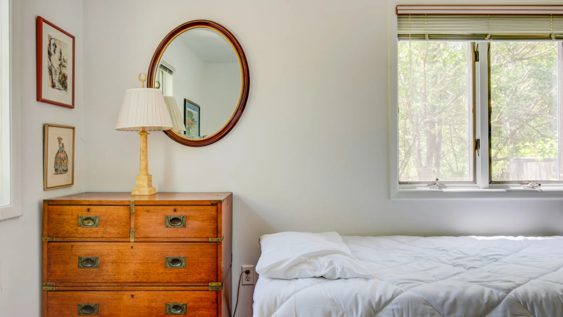 A bedroom with a wooden dresser beside a bed and a bright window.