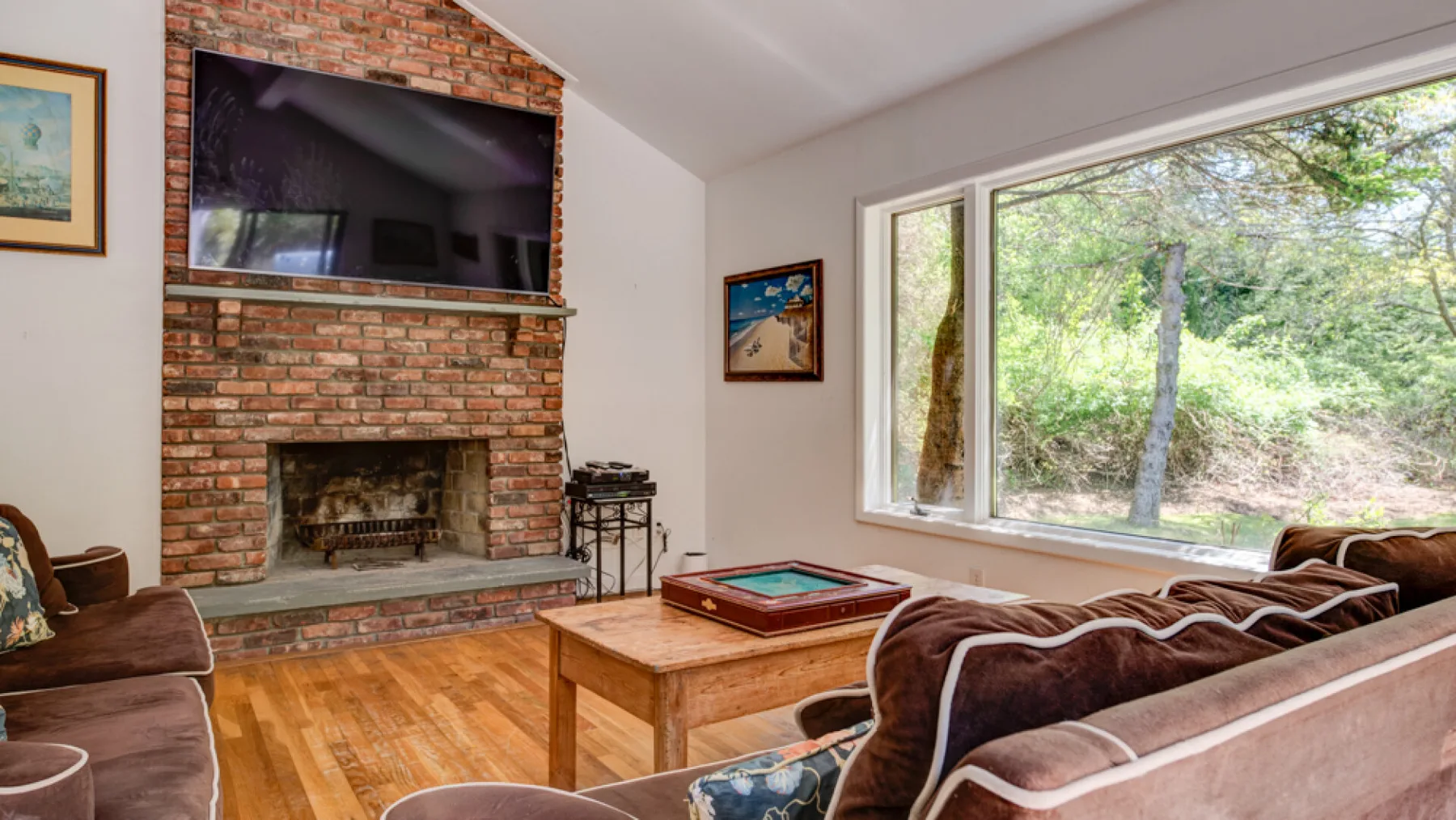 A living room has a brick fireplace, a television, and large windows overlooking trees.