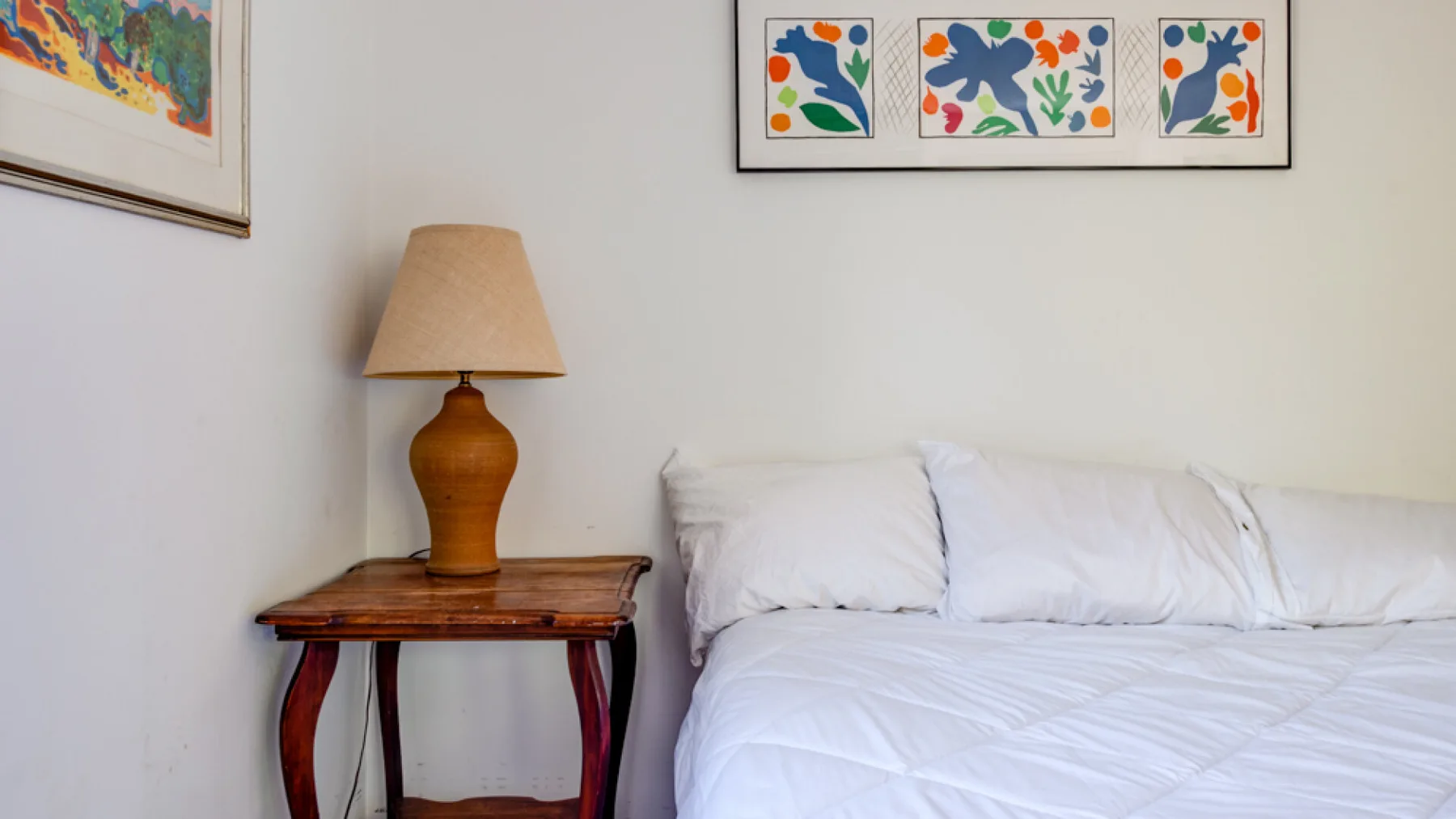 A bedroom with a bed, a wooden side table, and colorful framed prints.