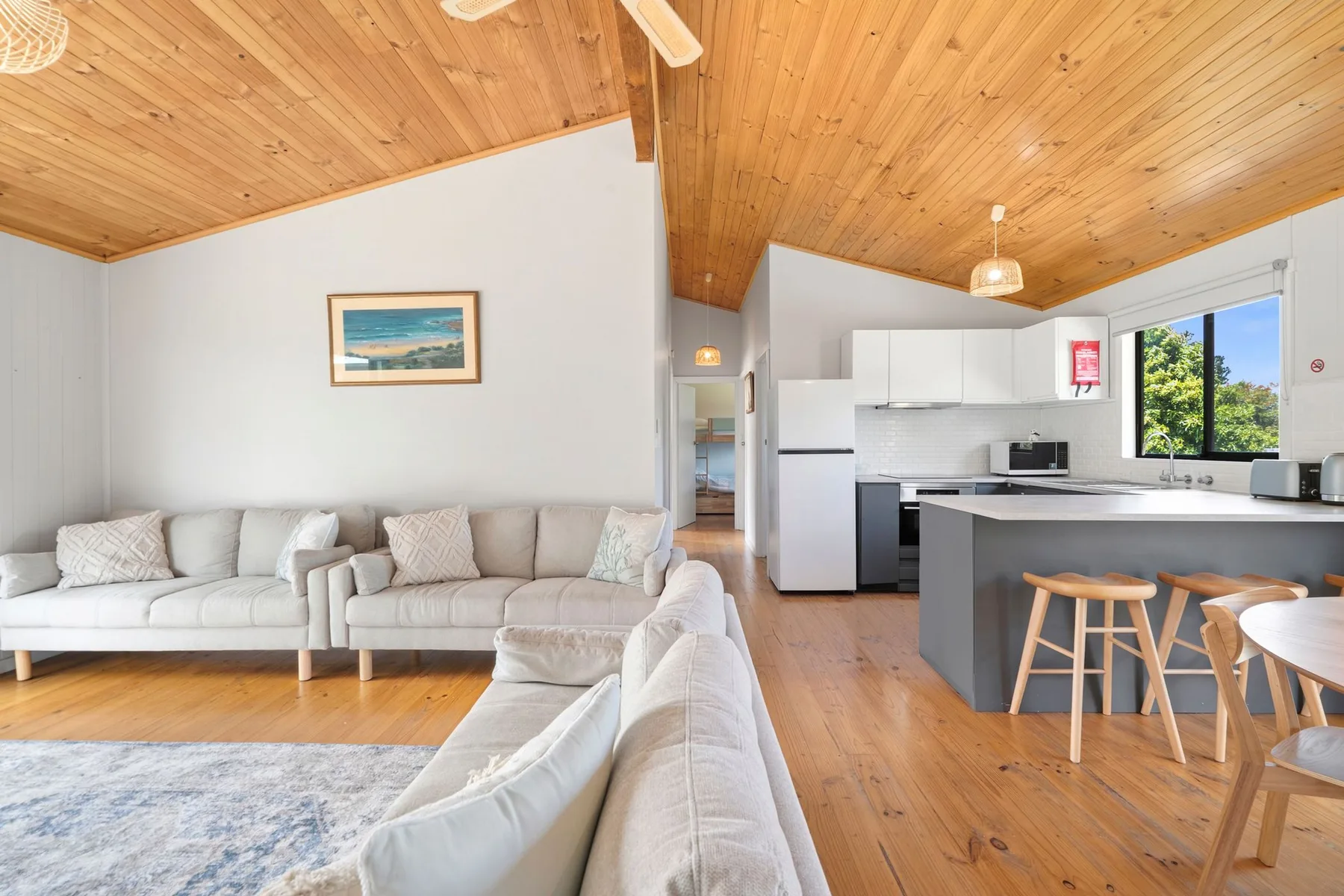 An open-plan living room and kitchen with pale seating and a wooden ceiling.