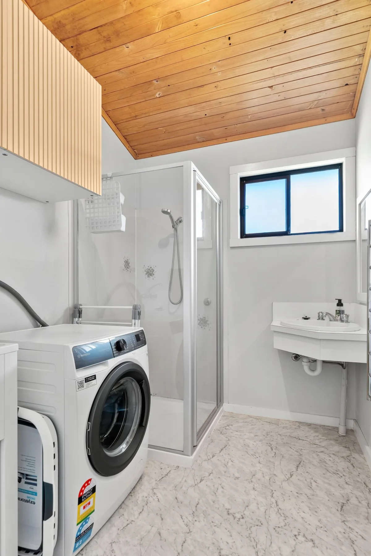 A bright bathroom with a shower, sink, and front-loading washing machine.