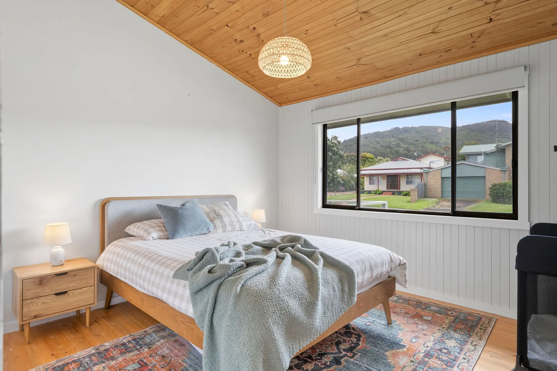 A bedroom with a bed beneath a wood-paneled ceiling and a large window.