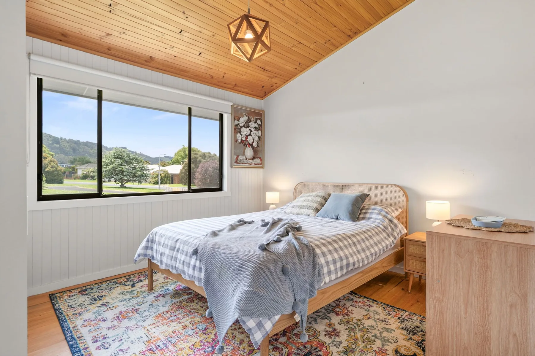 A bedroom with a bed beneath a wood-paneled ceiling and a large window.