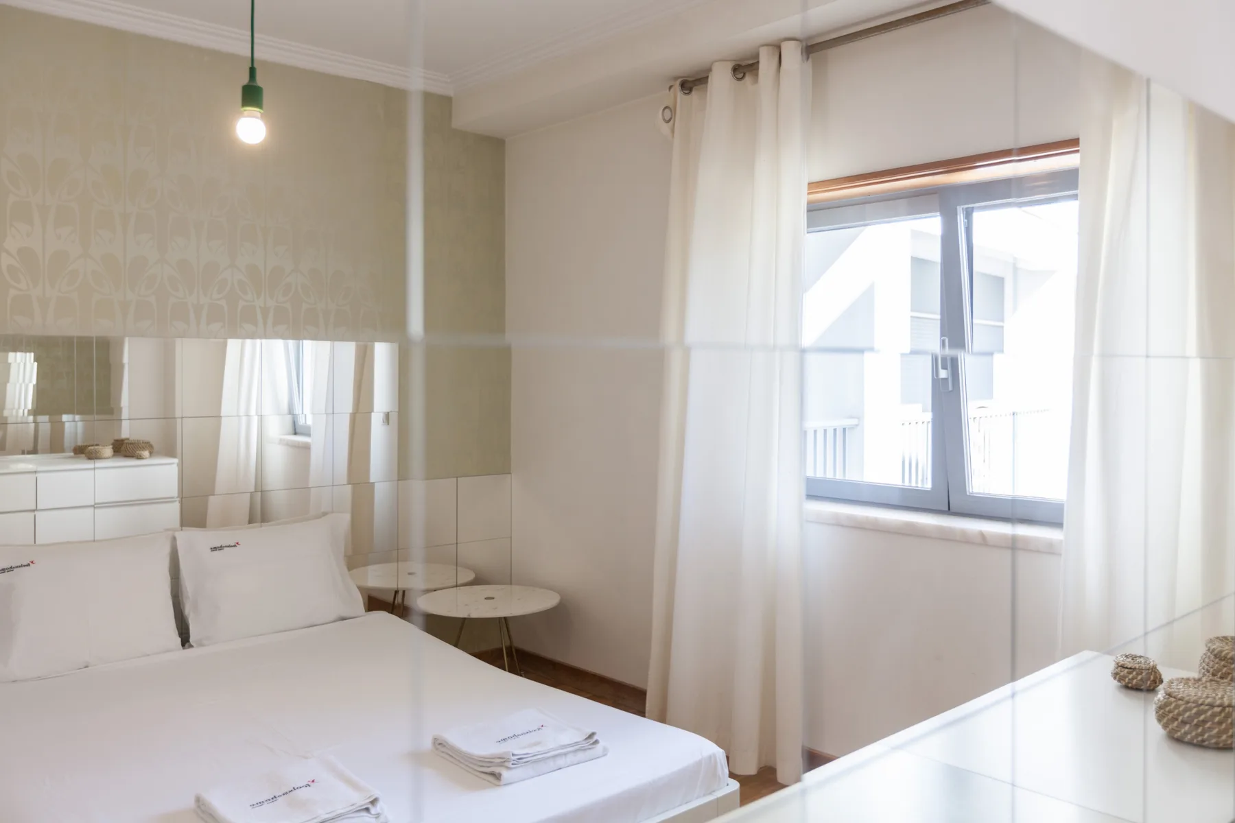 Un dormitorio luminoso con una cama, una pared con espejo y una gran ventana con cortinas.