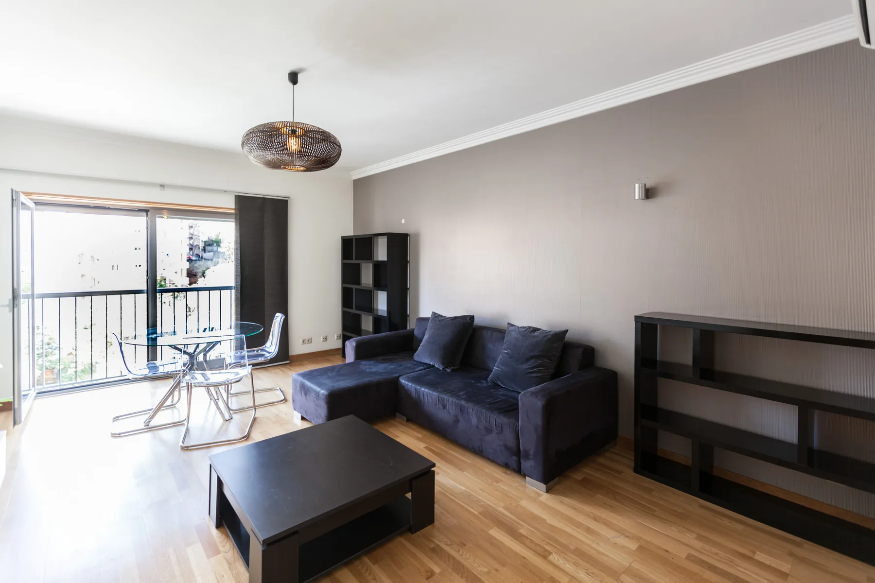 Sala de estar con asientos oscuros, una mesa de centro y una mesa de comedor de vidrio junto al balcón.