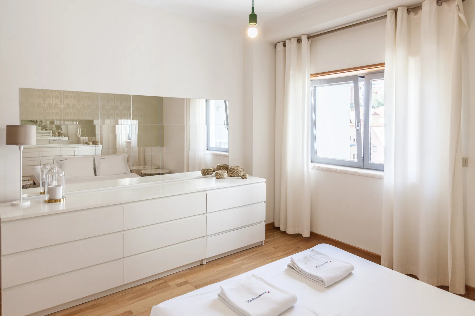 Un dormitorio luminoso con una cama blanca, una cómoda y una ventana con cortinas.