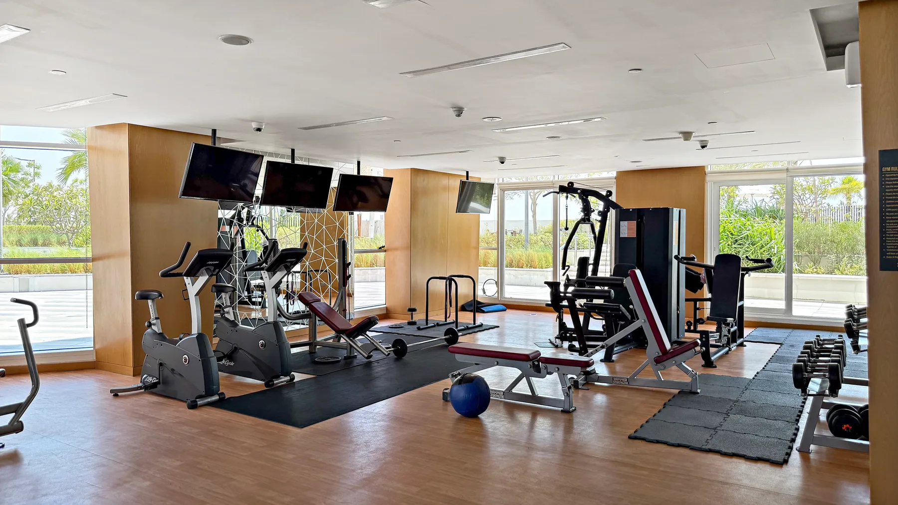 A bright indoor gym with exercise equipment and large windows overlooking greenery.
