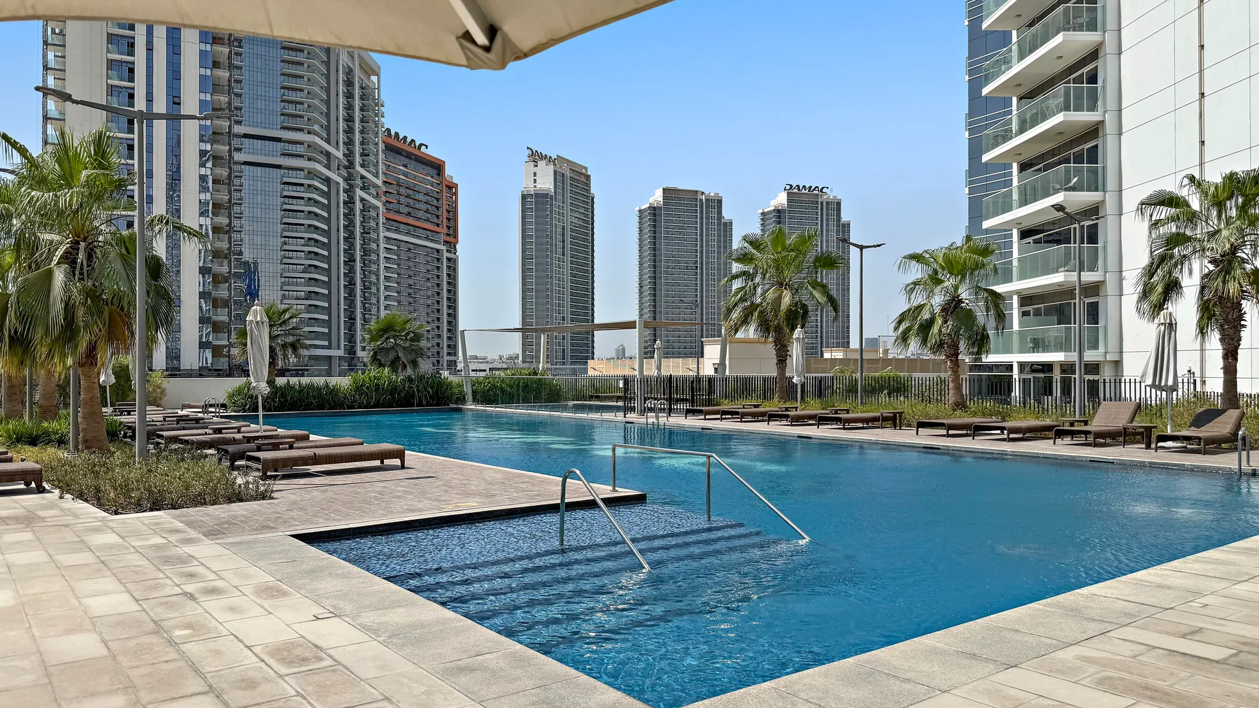 A swimming pool sits among high-rise buildings, with palm trees along its edge.