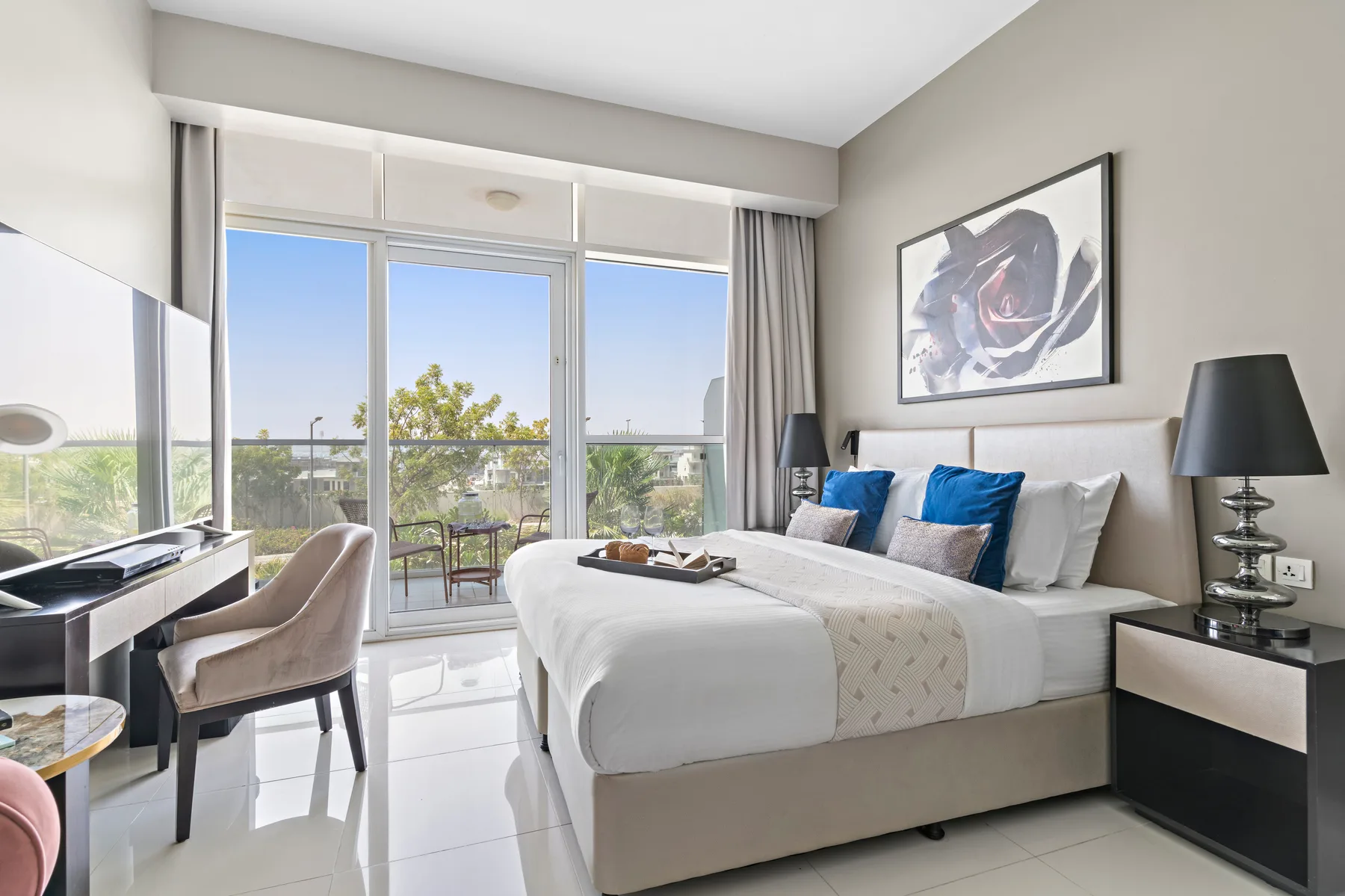 Un dormitorio luminoso con una cama grande, almohadas azules y ventanas con vistas a un balcón.