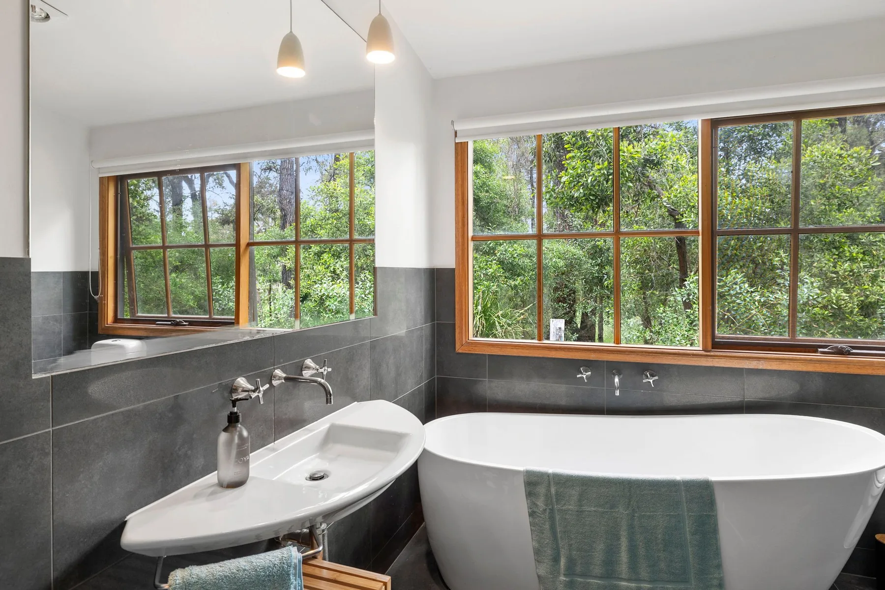 Un baño con bañera exenta, lavabo y grandes ventanas con vistas a la vegetación.