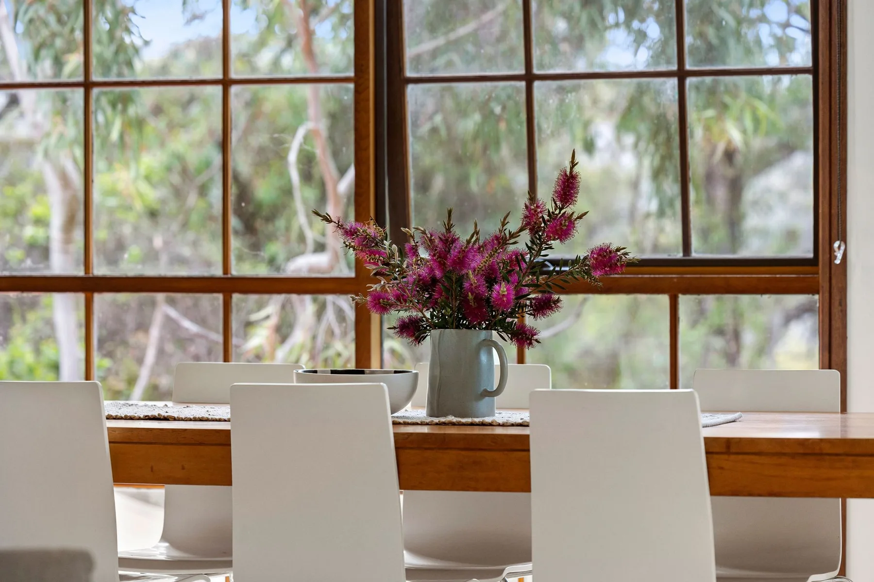 Una mesa de comedor con asientos blancos y flores está junto a grandes ventanas.