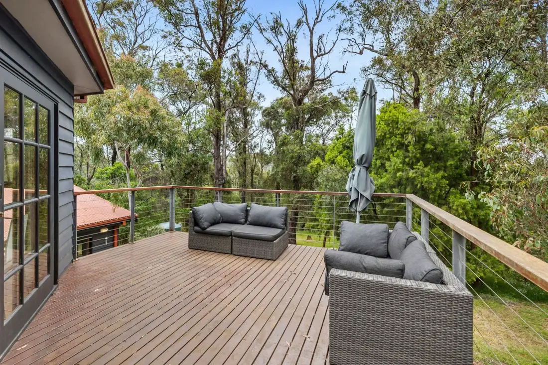 Una terraza de madera tiene asientos y una sombrilla cerrada, con árboles al otro lado de la barandilla.
