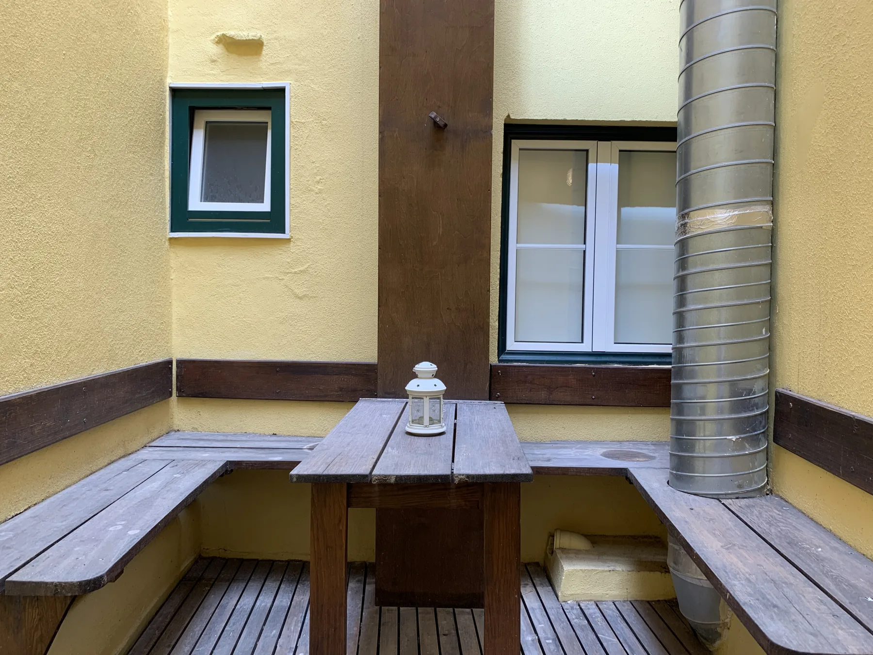 A small courtyard has wooden benches around a table beneath a tall metal pipe.
