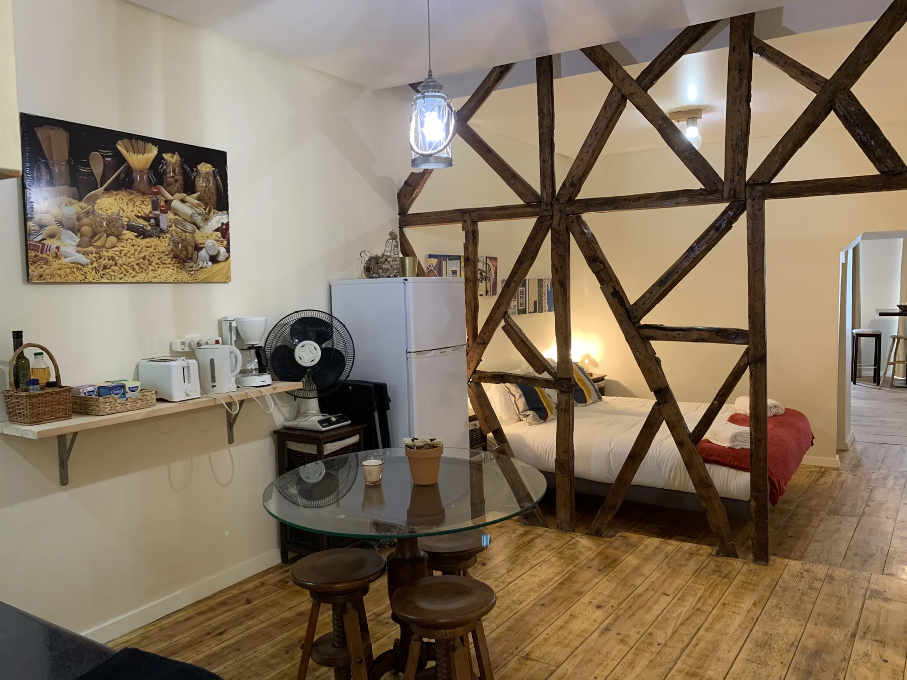 A bedroom with a bed behind a dark timber partition, with a small kitchen area nearby.