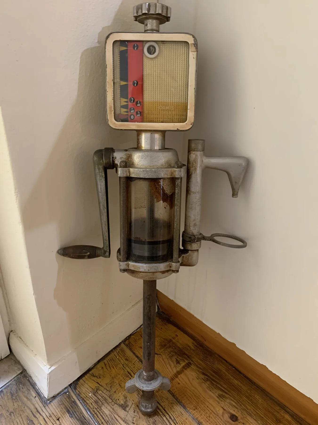 A vintage metal device stands beside a white wall on a wooden floor.
