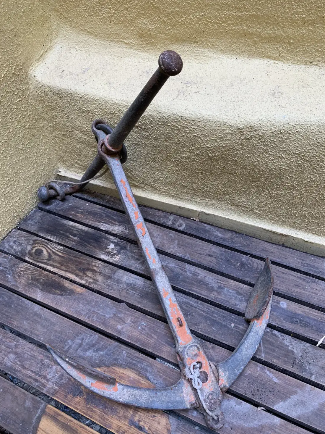 An old metal anchor rests on a dark wooden deck beside a textured wall.