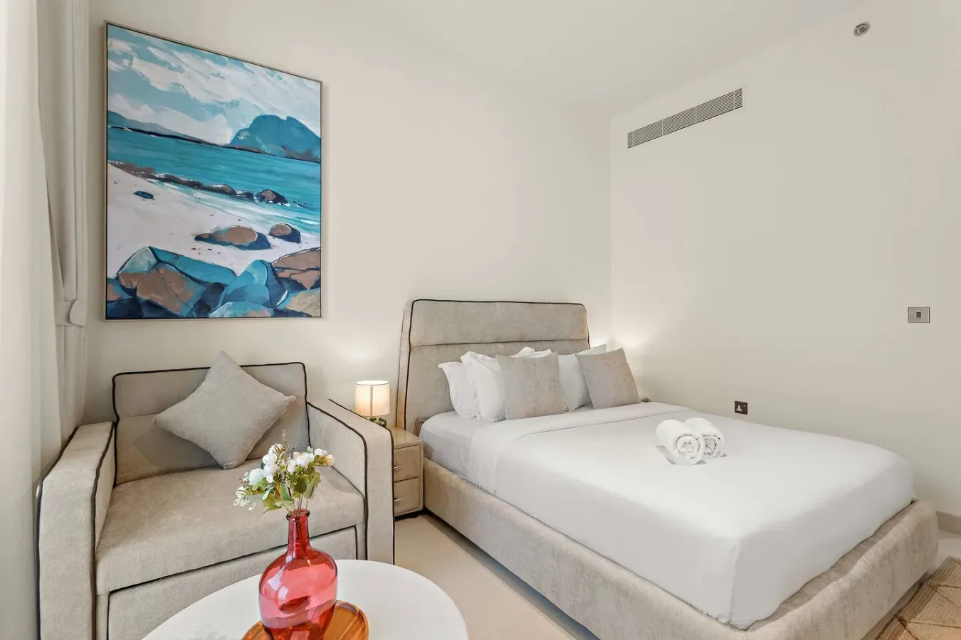 Un dormitorio luminoso con una cama bien arreglada, un sillón y arte costero.