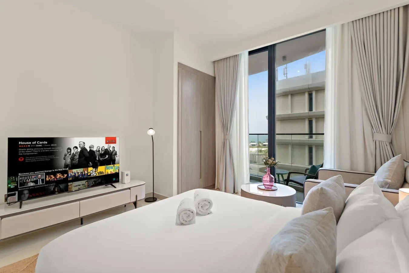Un dormitorio luminoso con una cama tendida, un televisor y grandes ventanas con cortinas.
