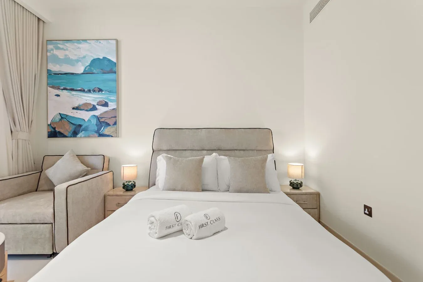 Un dormitorio con una cama tendida, mesitas de noche y un cuadro costero enmarcado.