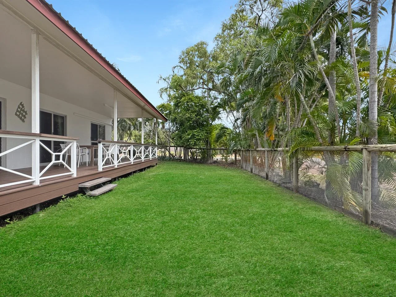 A grassy yard beside a covered veranda and a fence lined with tropical trees.