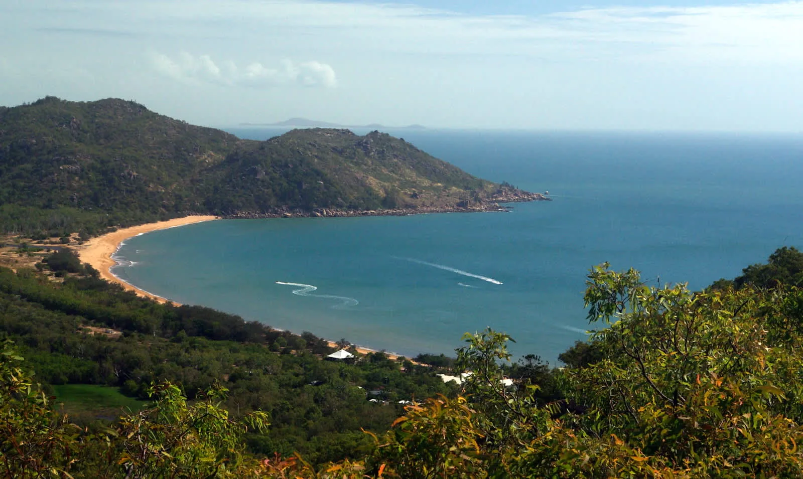 A curved sandy beach borders a blue bay beneath green hills.