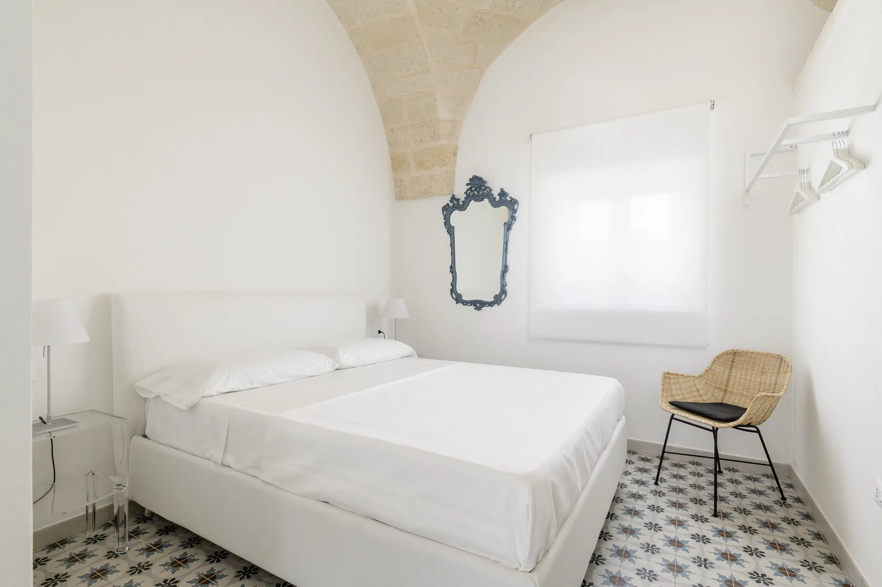 Un dormitorio luminoso con una cama blanca, suelo estampado y una silla junto a la ventana.