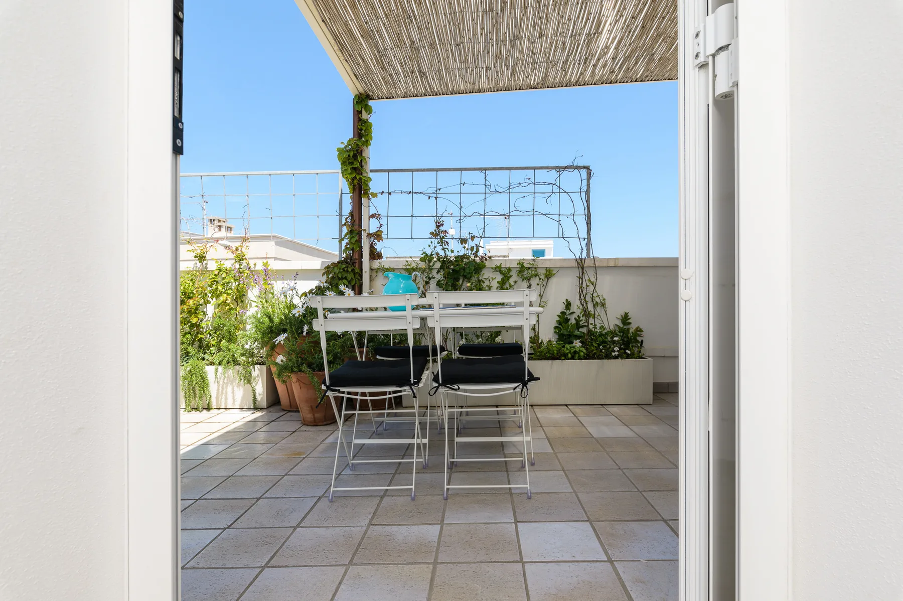 Una terraza exterior con suelo de baldosas, mesa, asientos y plantas en macetas.