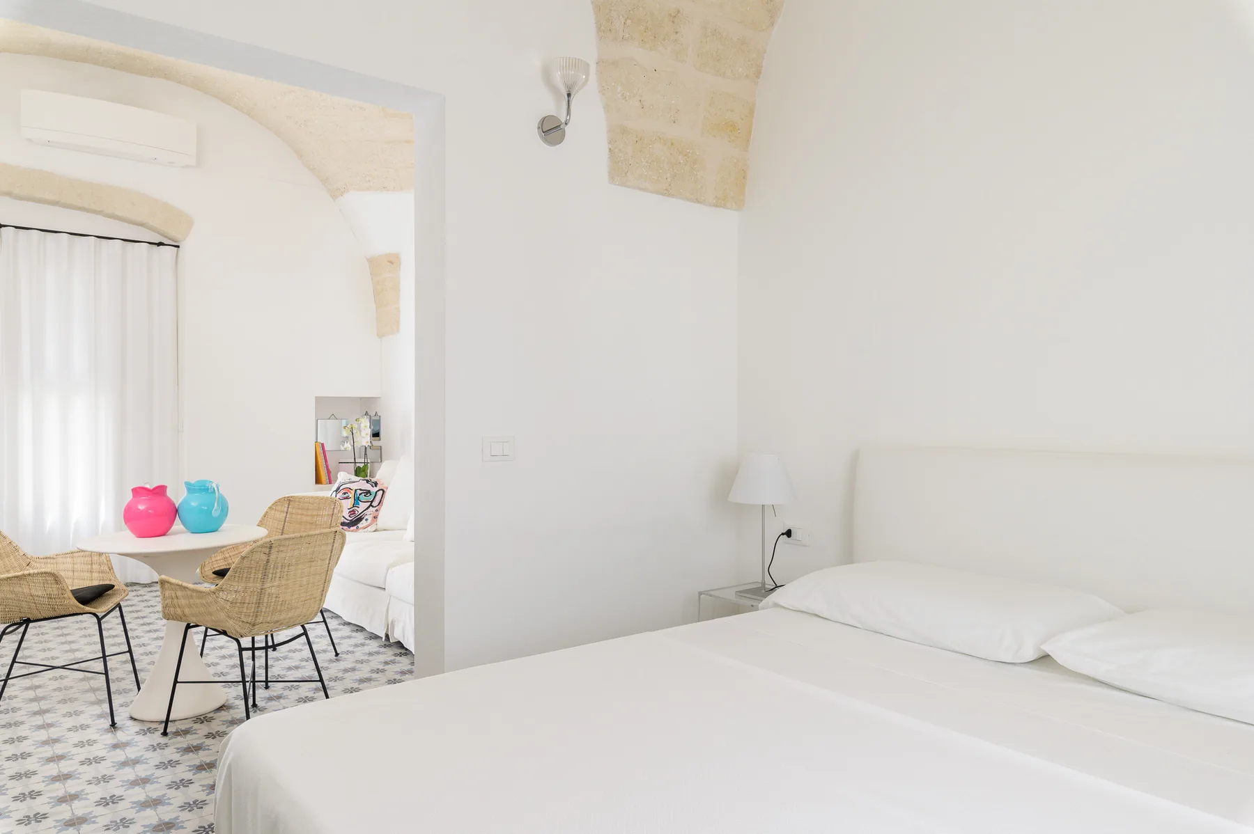 Un dormitorio luminoso con una cama blanca y una pequeña zona de comedor junto a la ventana.